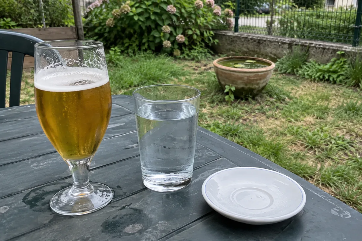 Dans un jardin communal en France, une table extérieure porte un verre de bière à côté d’un verre d’eau et d’une soucoupe, tandis qu’un petit pot rempli d’eau stagnante se trouve …