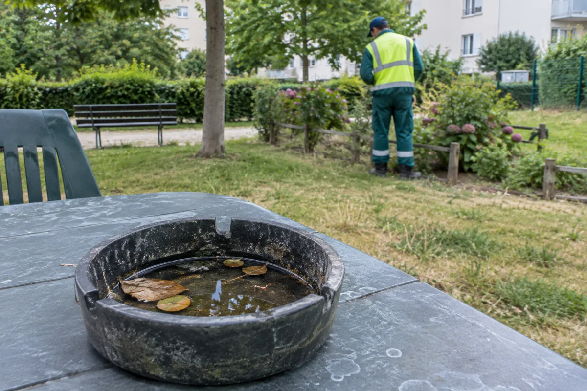 Dans un jardin communal en France, un cendrier posé sur une table de terrasse contient un peu d’eau stagnante et des feuilles, tandis qu’un agent municipal de dos l’examine à dist…