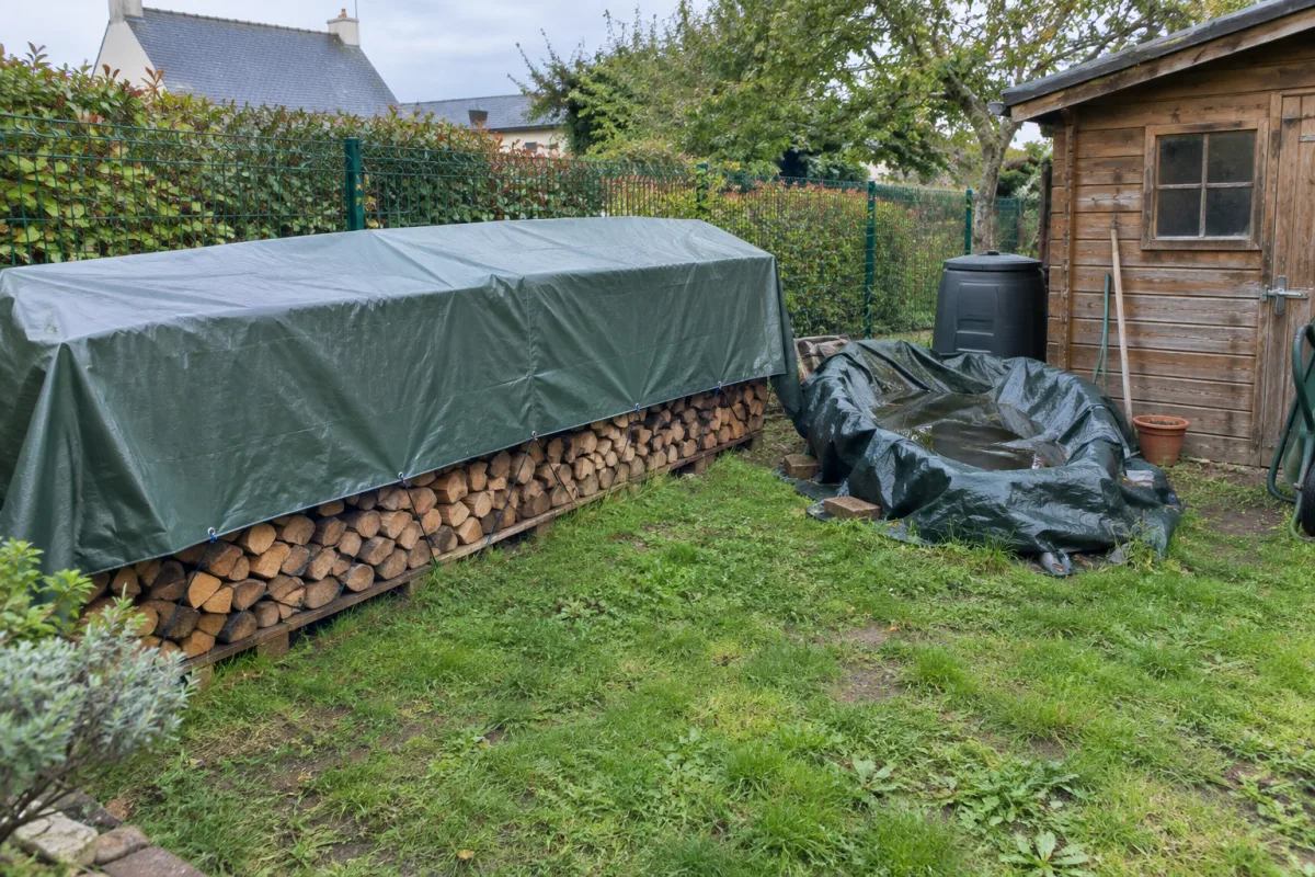 Dans un jardin d’une commune française, une bâche soigneusement tendue couvre du bois tandis qu’une autre, mal repliée, retient un peu d’eau de pluie dans ses plis près d’un abri …