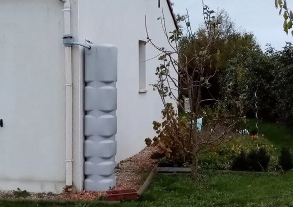Récupérateur d’eau de pluie gris branché sur la descente de gouttière, contre le mur d’une maison